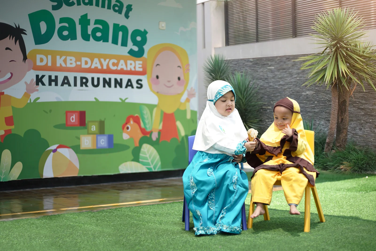 Dua anak duduk bersama di area luar dengan tanaman